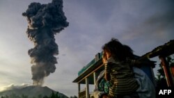 A woman and child look on at volcanic ash rising into the air during the eruption of Mount Ibu, as seen from Duono village, North Maluku province, Jan. 15, 2025. 